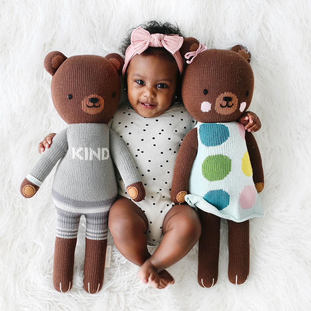 A smiling baby with their arms around two cuddle+kind stuffed dolls, Oliver and Ivy the bears.