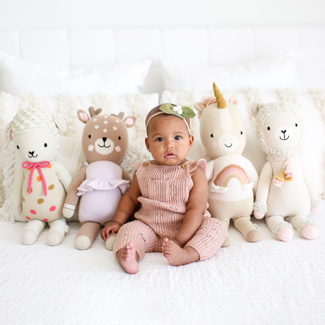 A baby, sitting on a bed, in the middle of four cuddle+kind stuffed dolls.