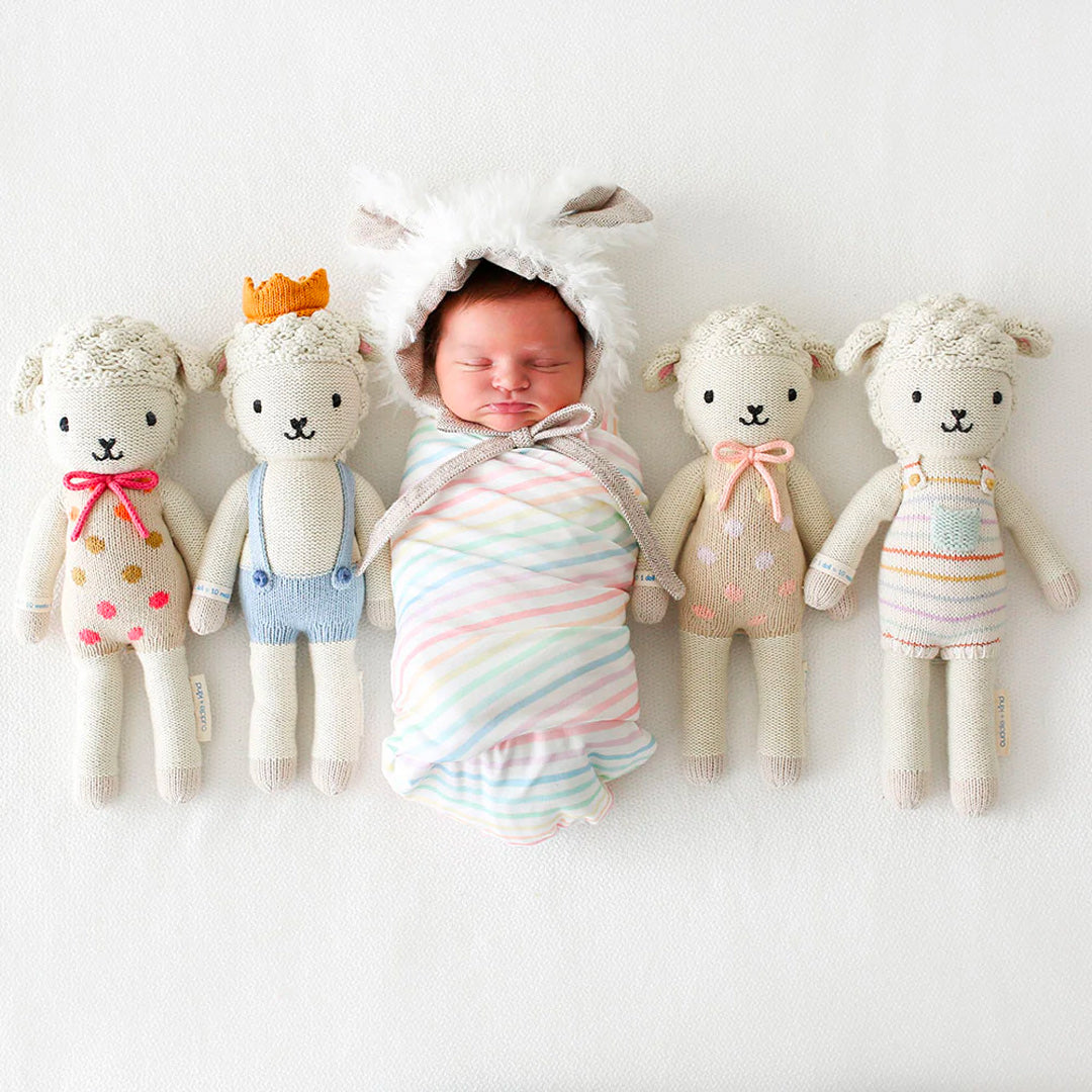 A sleeping baby in a lamb hood with four stuffed lamb dolls: Lucy the lamb, Sebastian the lamb, Lucy the lamb in pastel, and Avery the lamb.