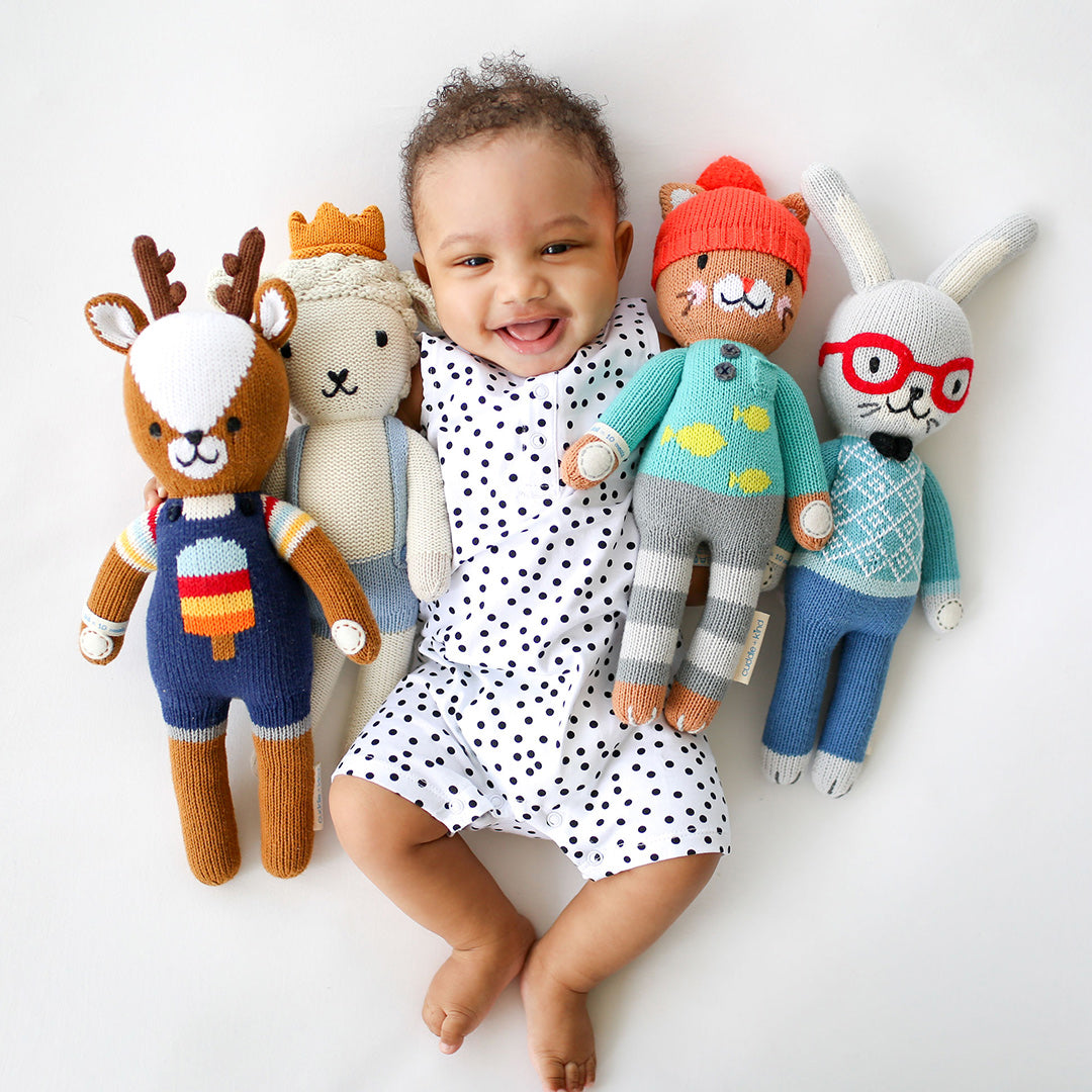 A laughing baby with his arm around several cuddle and kind stuffed dolls.