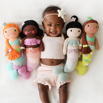 A laughing baby holding four mermaid stuffed dolls: Isla, Maya, Luna and Pearl.