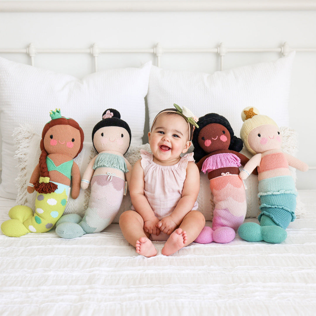 A laughing baby sitting between four mermaid stuffed dolls: Skye, Isla, Maya and Pearl.