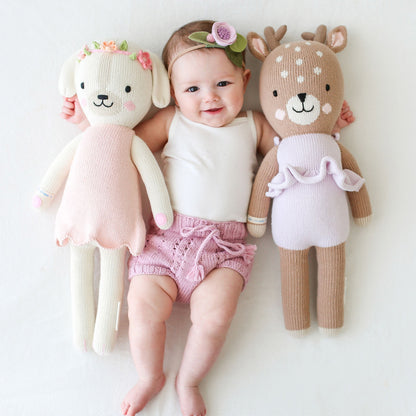 A smiling baby wearing a floral headband with their arms around a Charlotte the dog stuffed doll and a Violet the fawn stuffed doll.