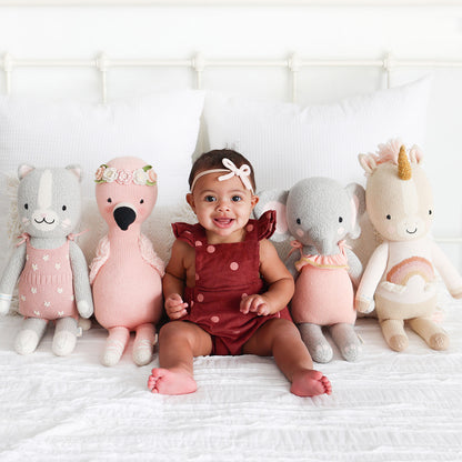 A smiling baby sitting on a bed in the middle of four cuddle+kind stuffed dolls: Daisy, Penelope, Eloise and Zara.