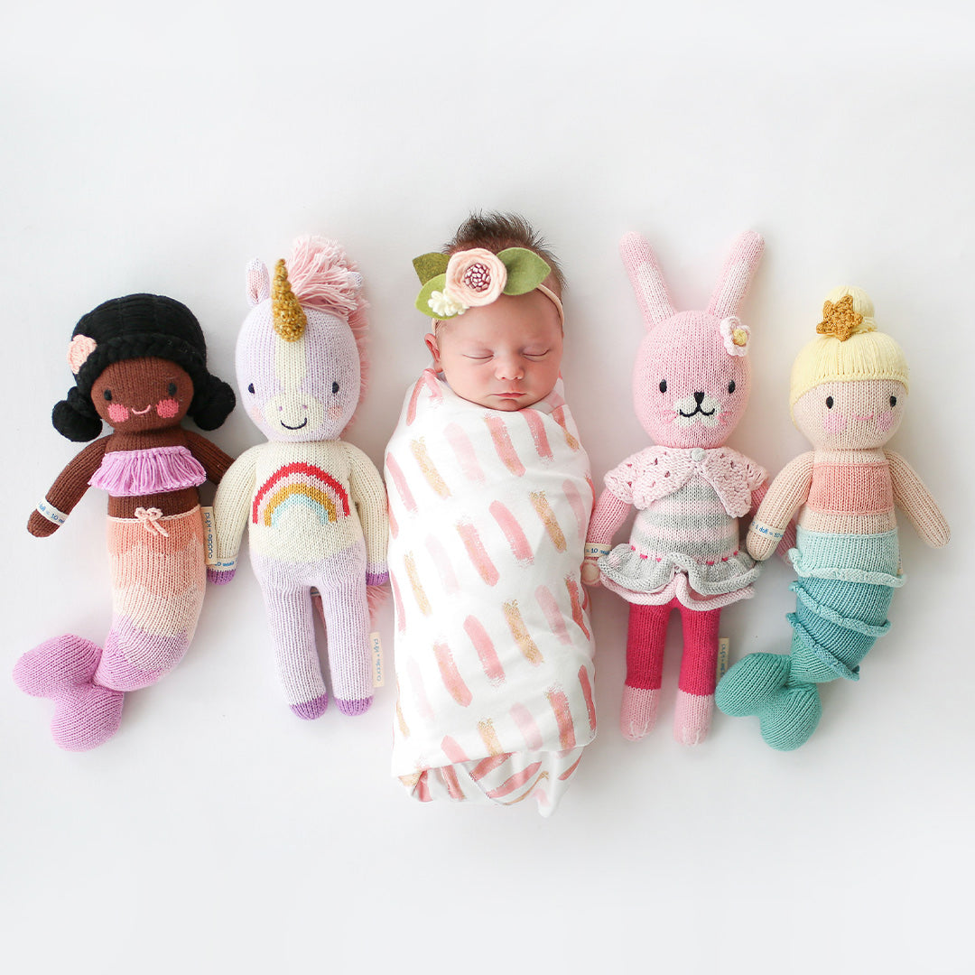 A swaddled, sleeping baby lying in the middle of four cuddle+kind stuffed dolls: Maya, Zoe, Chloe and Skye.