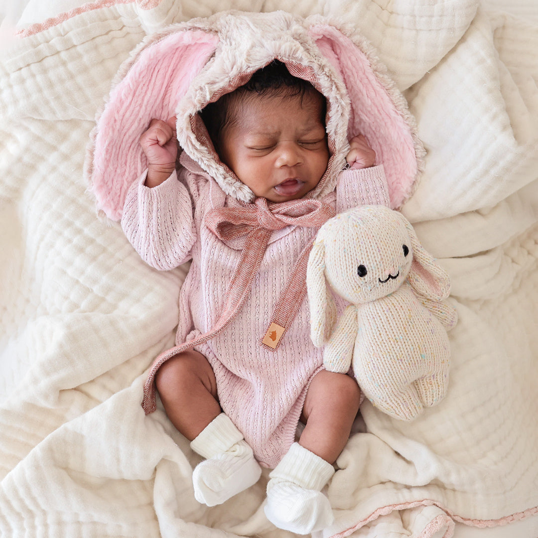 A sleeping baby in a bunny-eared bonnet beside a baby bunny in confetti.