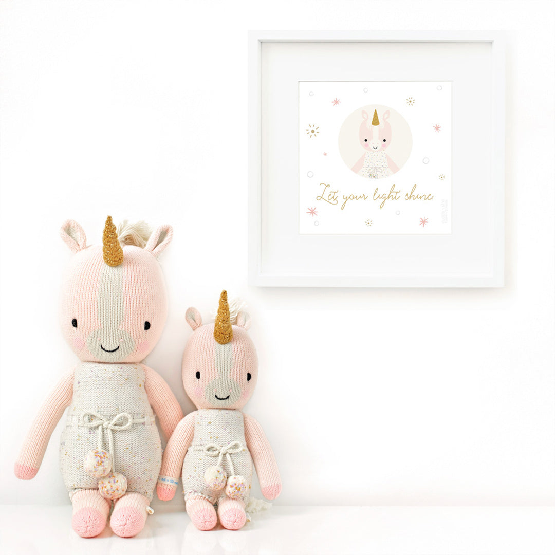 Two Ella the unicorn stuffed animals sitting beside a framed print with a picture of Ella that says “Let your light shine.”