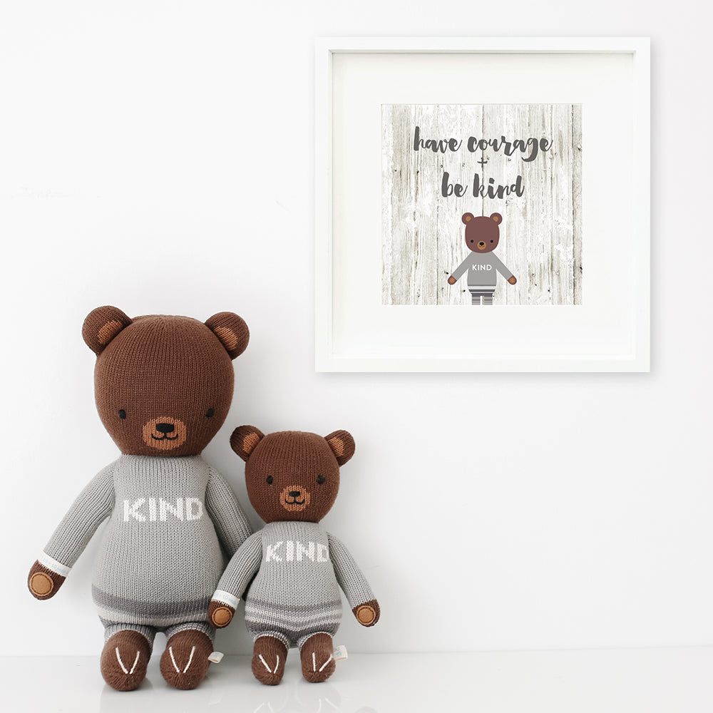 Two Oliver the bear stuffed animals sitting beside a framed print with a picture of Oliver that says “Have courage + be kind.”