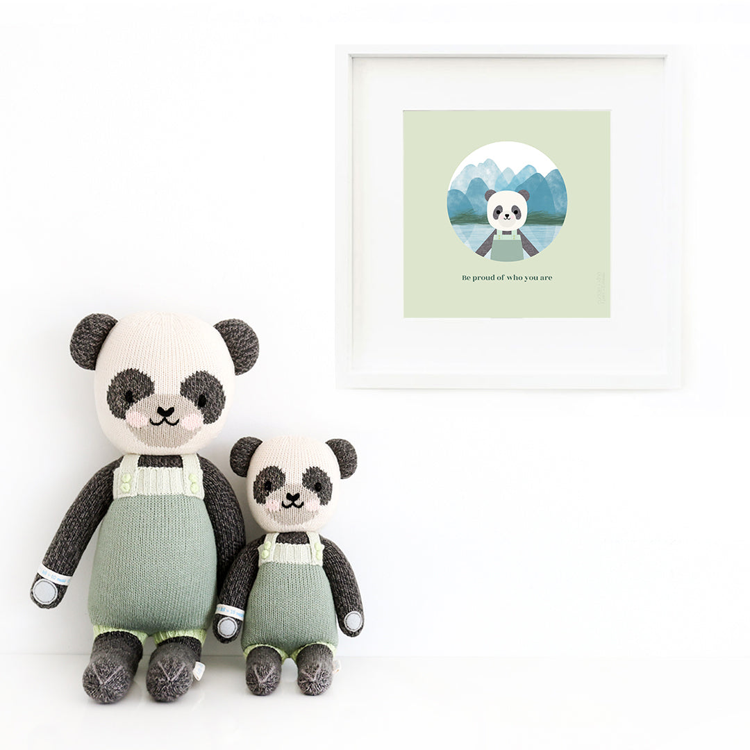 Two Paxton the panda stuffed dolls sitting beside a framed print with a picture of Paxton, with text that says "Be proud of who you are."