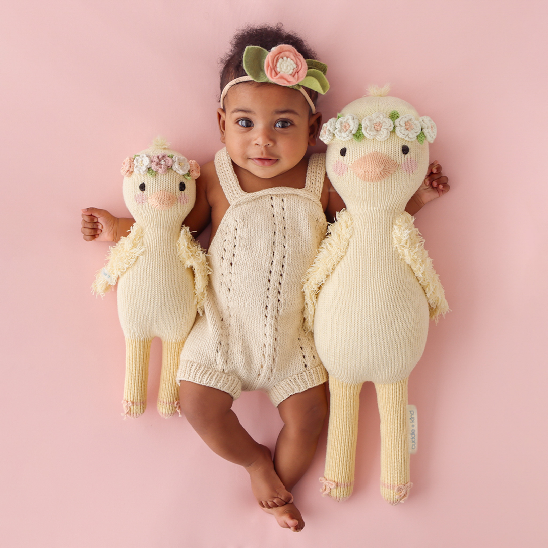 A smiling baby, wearing a floral headband, with their arms around two Flora the duckling stuffed dolls, in the regular and little size.