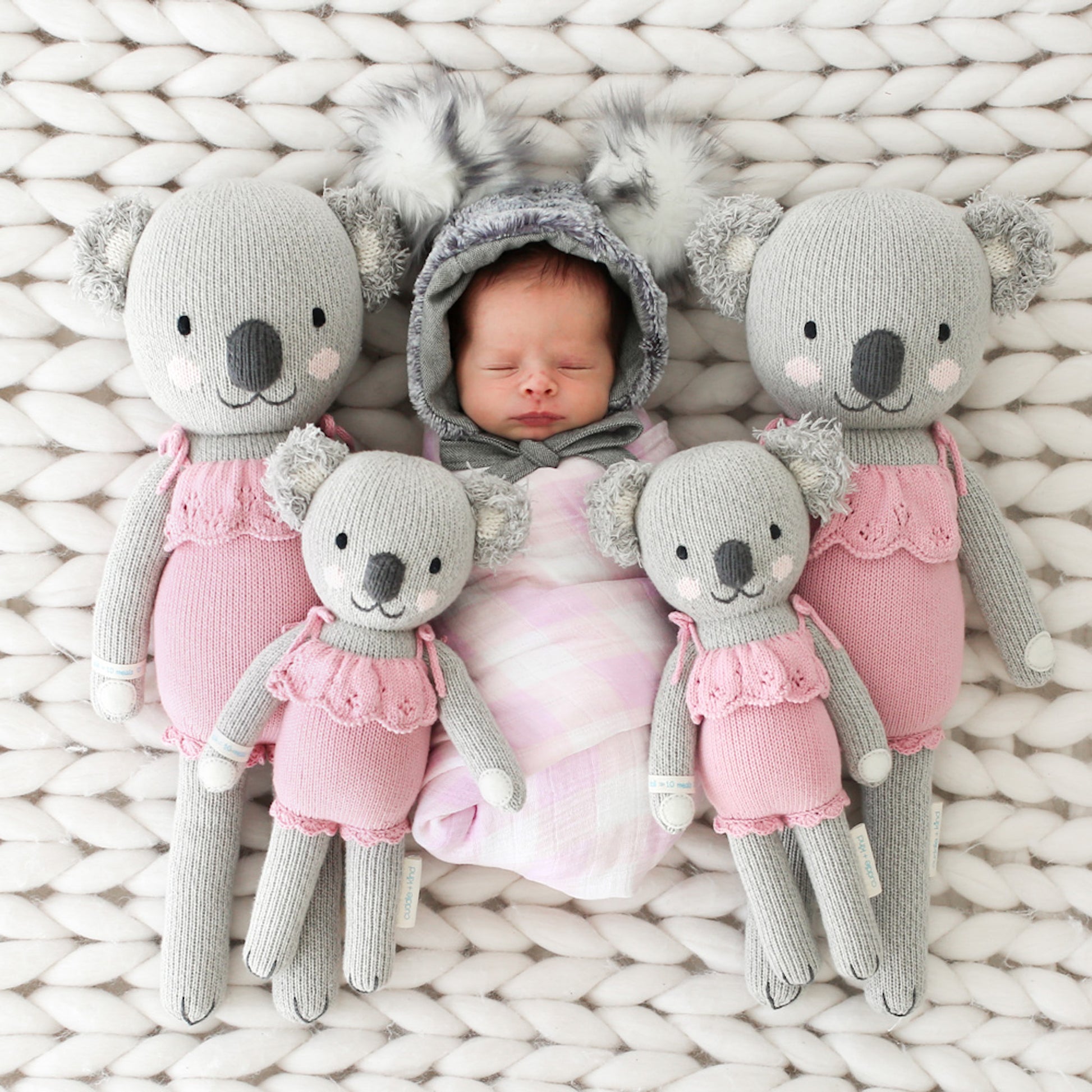 A baby in a koala hood lying in between four Claire the koala dolls in the little and regular sizes.