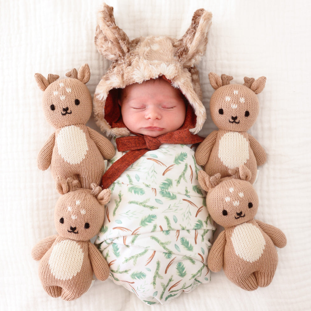 A sleeping baby in a fur bonnet nestled alongside 4 baby fawn stuffed dolls.