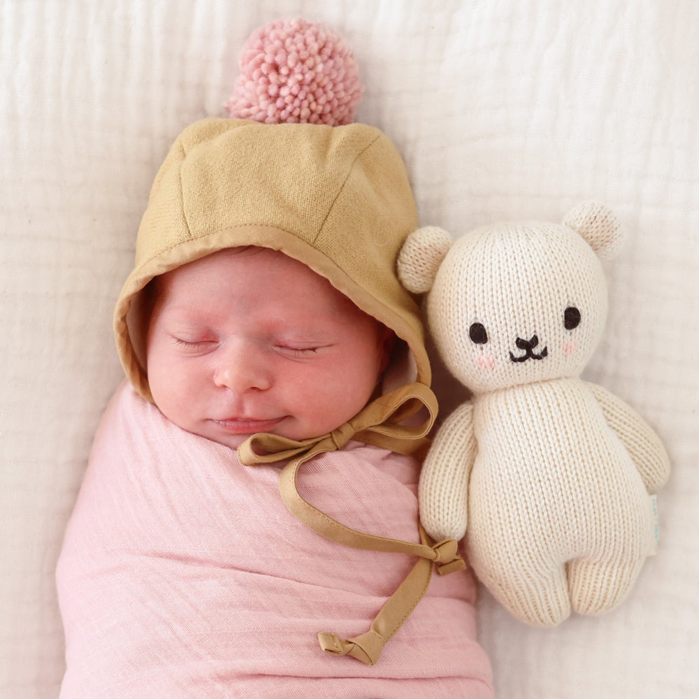 A sleeping baby, wearing a pom-pom bonnet, snuggled next to a baby polar bear.