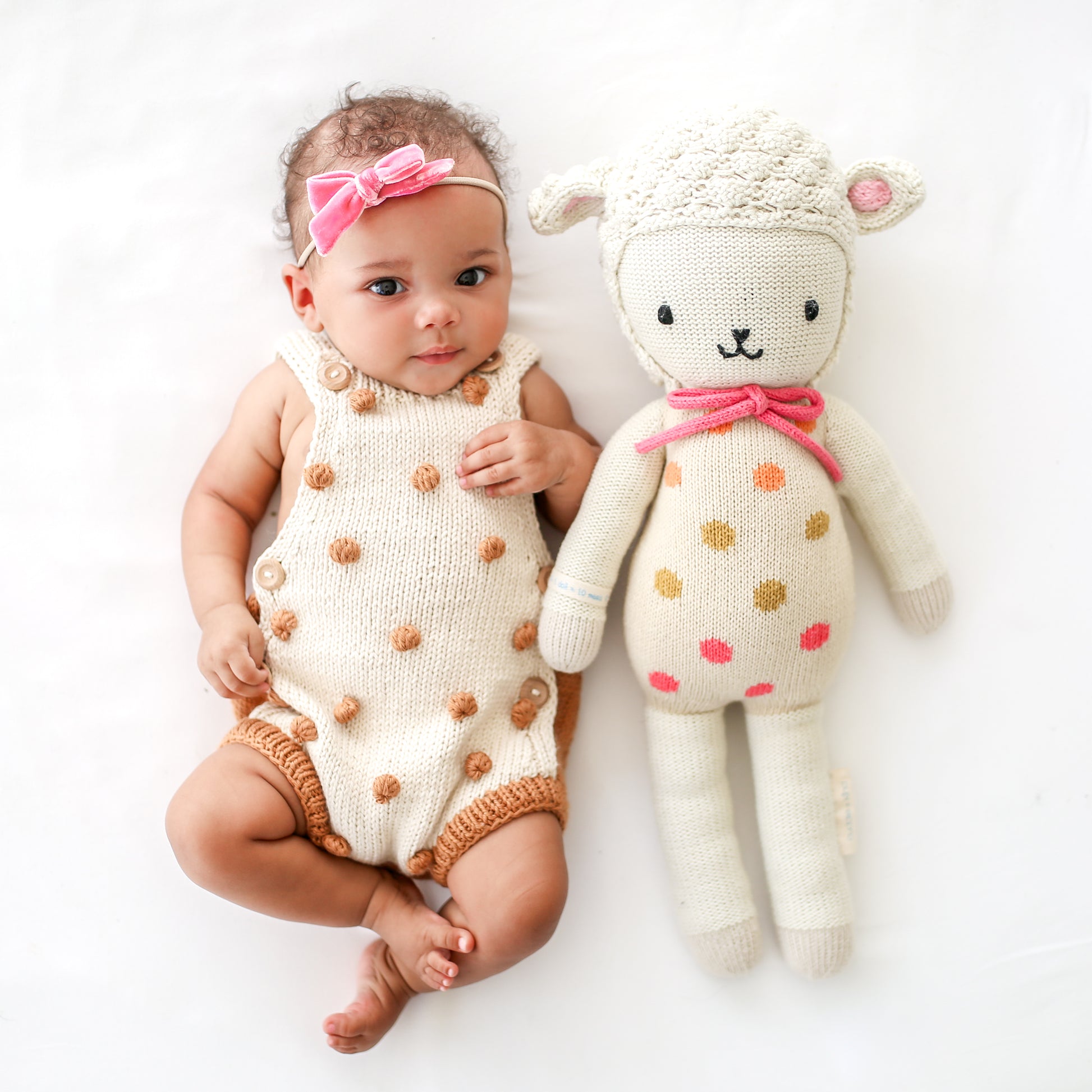 A baby wearing a knit romper and a pink, bowed headband, lying beside Lucy the lamb.