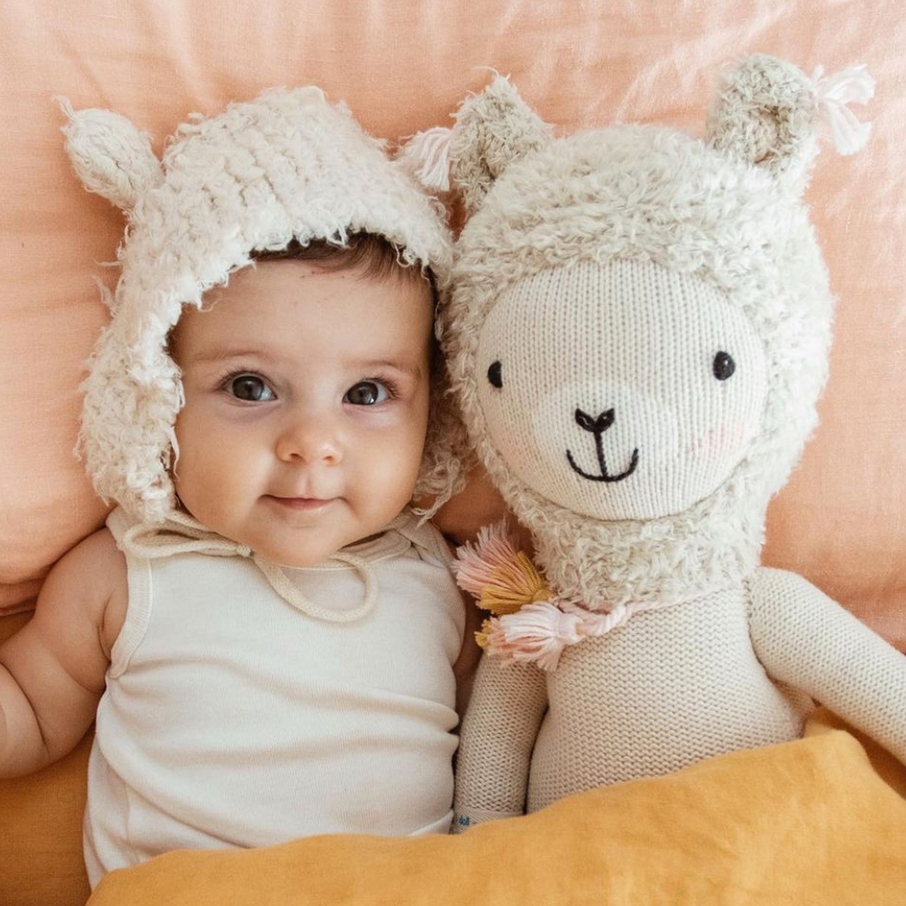 A baby, wearing a fuzzy, llama-eared bonnet, under a blanket with Lola the llama.